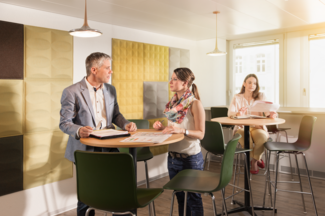 Mitarbeitende in gut gelüfteten Aufenthalsraum an einem Hochtisch in einer Besprechung. 